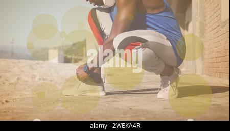 Taches jaunes de lumière contre la section médiane de l'homme afro-américain fit attacher des lacets de chaussures à la plage Banque D'Images