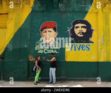 Un couple cubain socialisant devant un bâtiment peint avec les portraits de Hugo Chávez et Che Guevara dans la vieille Havane, Cuba. Banque D'Images