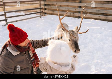 Vue arrière du petit garçon d'enfant et père aimant nourrissant mignon jeune renne sur la ferme enneigée le jour ensoleillé d'hiver. Banque D'Images