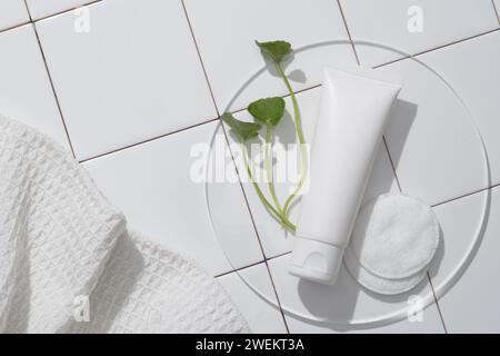 Maquette d'un produit cosmétique - nettoyant pour le visage avec un tube en plastique blanc non étiqueté, des feuilles de gotu kola sur un podium clair et un tampon de coton contre un dos Banque D'Images