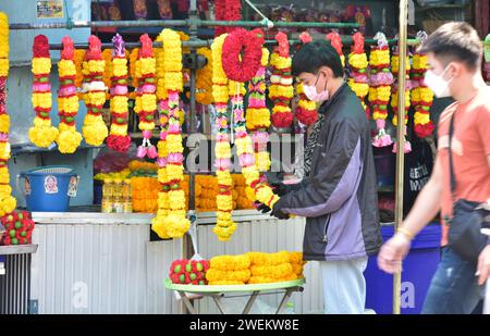 Un homme ajuste les guirlandes de fleurs qu'il vend depuis un étal à côté du temple Sri Maha Mariamman à Bangkok, Thaïlande, Asie du Sud-est. Les guirlandes de fleurs sont vendues comme offrandes pour la cérémonie hindoue dans le temple. Un homme portant un masque passe devant. Banque D'Images