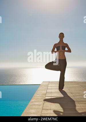 Young woman practicing yoga par une piscine avec l'océan dans l'arrière-plan Banque D'Images