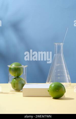 Citrons verts frais sur fond bleu avec verrerie de laboratoire et podium vide blanc. Espace pour l'affichage cosmétique de l'extrait de chaux. L'huile essentielle de ligne est idéale pour Banque D'Images