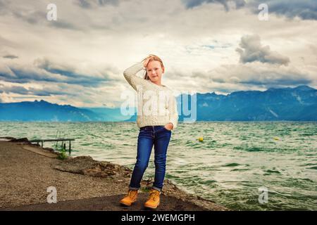 Mignonne petite fille de 8 ans jouant au bord du lac par une journée très venteuse, portant un pull blanc chaud tricoté, bras grands ouverts, image tonique Banque D'Images