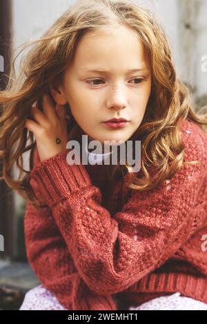 Gros plan portrait d'une mignonne petite fille de 8 ans avec les cheveux bouclés, portant un pull en terre cuite, assis sur fond de bois gris. Banque D'Images