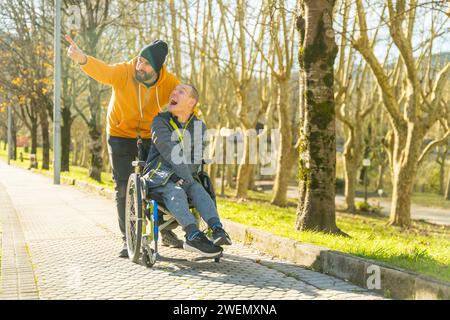 Homme handicapé assis en fauteuil roulant et ami appréciant la nature pointant et regardant autour dans un parc Banque D'Images