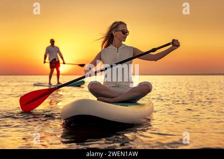 Jeune homme et femme marche avec des planches sup au lac Sunset. Belle fille est assise sur stand up paddle board au lac calme Banque D'Images