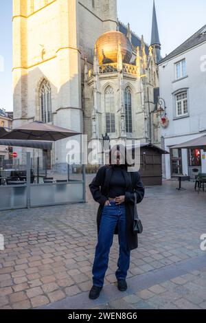 Cette photographie montre une jeune femme en tenue à la mode, debout avec confiance sur une vieille place européenne avec une architecture historique en arrière-plan. Son style moderne contraste avec la pierre ancienne et la maçonnerie détaillée de la cathédrale derrière elle, illustrant un mélange du présent et du passé. La place vide évoque un moment tranquille d'exploration ou de réflexion au milieu d'un environnement typiquement animé. Exploration élégante dans un cadre historique. Photo de haute qualité Banque D'Images