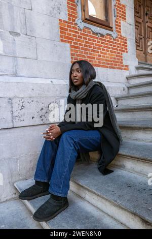 Cette image capture une jeune femme afro-américaine assise sur les marches en pierre d’un bâtiment urbain, son expression réfléchie suggérant un moment de calme dans une ville animée. Le contraste de sa tenue moderne et décontractée avec l'architecture classique qui l'entoure crée un récit visuel dynamique. Elle semble à l’aise dans l’environnement urbain, sa présence mêlant vie contemporaine et structures urbaines intemporelles. C'est un instantané de l'individualité et des espaces personnels que nous sculptons dans les paysages urbains. Jeune femme sur City Steps. Photo de haute qualité Banque D'Images