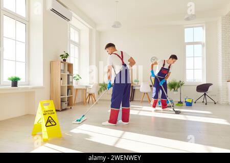Jeune équipe de concierges en uniforme laver le sol avec signe de prudence sol mouillé à la maison. Banque D'Images