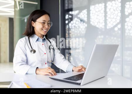 Femme médecin travaillant à l'ordinateur portable tout en étant assis au bureau et souriant dans le bureau de l'hôpital. Femme médicale asiatique fournissant une consultation en ligne sur ordinateur portable et se sentant heureuse après avoir entendu de bonnes nouvelles. Banque D'Images