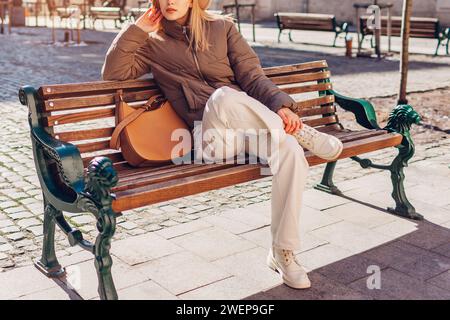 Jeune femme à la mode assise sur un banc dans la rue de la ville européenne portant une veste élégante, un pantalon et des bottes par sac à main. Tenue de printemps. Beige blanc et b Banque D'Images