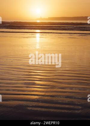 Coucher de soleil alors que les vagues roulent vers le sable humide ondulé à motifs avec une île derrière sur une plage à Essaouira, Maroc, le 26 janvier 2024 Banque D'Images