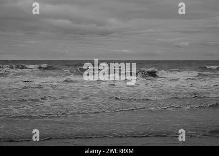 Les vagues s'écrasent sur la plage créant de l'écume blanche sous les nuages d'un ciel sombre en monochrome. Banque D'Images