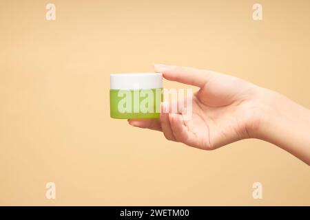 La main de la femme tient un pot de crème hydratante en verre ambré sur fond beige. Bouteille sans étiquette avec masque anti-âge, cosmétique. Concept de Banque D'Images