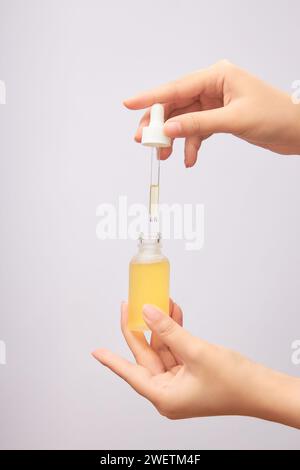 Bouteille en verre de sérum avec bouchon compte-gouttes, contenant de l'essence jaune dans les mains des femmes sur fond clair. Photo publicitaire pour produit cosmétique, skinca Banque D'Images