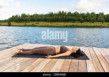 Une femme mince effectue un exercice relaxant shavasana après une séance d'entraînement principale, pratique le yoga dans des leggings et un haut court, se trouve sur un tapis sur le rivage de Banque D'Images