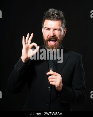 Jeune homme portant l'uniforme de prêtre debout sur fond blanc faisant geste ok avec la main souriante, oeil regardant à travers les doigts avec le visage heureux. Banque D'Images