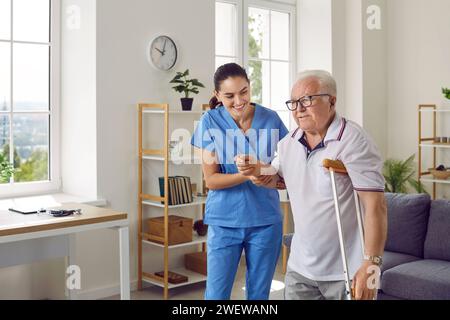 Infirmière souriante tenant son patient âgé par la main et l'aidant à marcher avec une béquille Banque D'Images