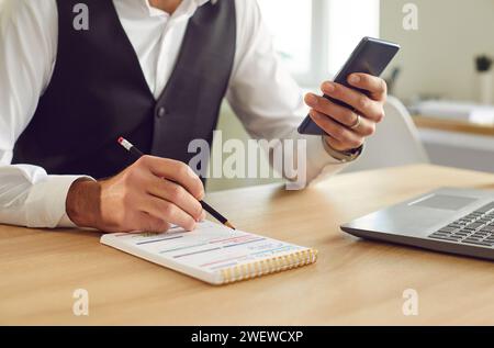Homme d'affaires écrivant pour faire la liste dans le bloc-notes assis au bureau avec ordinateur portable avec smartphone à la main. Banque D'Images