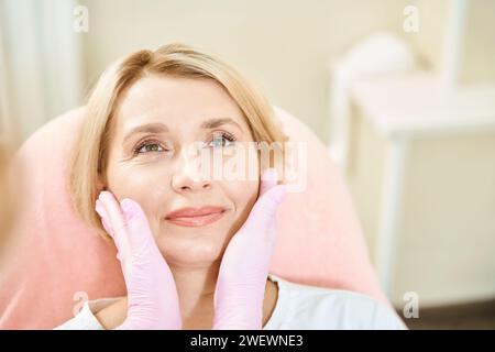 Mains de cosmétologue féminin recadrées examinant et touchant le visage d'une femme souriante Banque D'Images