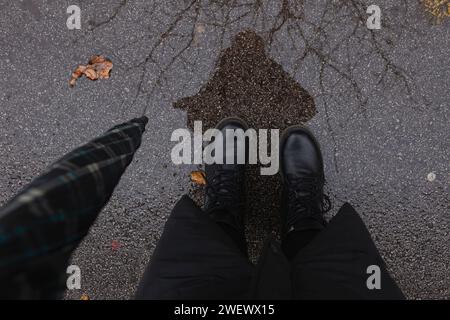 Vue de dessus des pieds de la femme en bottes d'hiver noires debout sous la pluie sur la rue humide, tenant un parapluie Banque D'Images