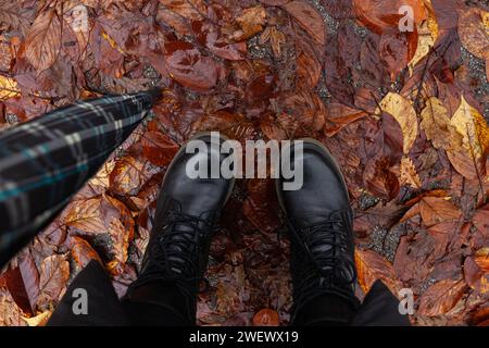 Vue de dessus des pieds de la femme en bottes d'hiver noires debout sous la pluie sur la rue humide en pile de feuilles tombées, tenant un parapluie Banque D'Images