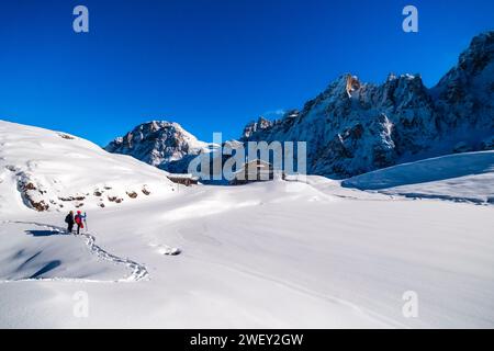 Deux randonneurs en raquettes marchent vers la cabane de montagne Baita Segantini, les sommets du groupe Pale au loin, en hiver. Banque D'Images