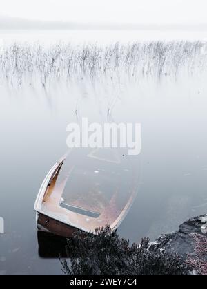 Une scène sereine mettant en vedette un bateau partiellement submergé reposant dans les eaux calmes d'un lac brumeux, suggérant le silence tranquille de l'aube. Banque D'Images