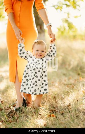 Grande fille souriante fait ses premiers pas avec l'aide de sa mère à l'extérieur. Banque D'Images