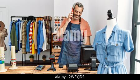 Homme d'âge moyen à cheveux gris assistant de magasin utilisant l'ordinateur parlant sur le smartphone à la boutique de vêtements Banque D'Images