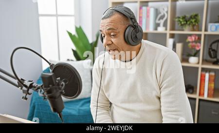 Un homme hispanique d'âge moyen parle dans un micro tout en portant un casque dans un studio de radio moderne. Banque D'Images