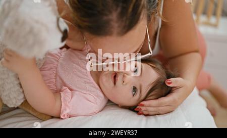 Dans le confort de leur maison, mère et fille partagent une matinée relaxante au lit, souriant et exprimant leur amour en embrassant de manière ludique l’ours en peluche i. Banque D'Images