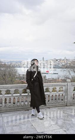 Une femme souriante vêtue d'un manteau noir pose avec la ligne d'horizon d'istanbul en arrière-plan, capturant les voyages et l'exploration culturelle. Banque D'Images