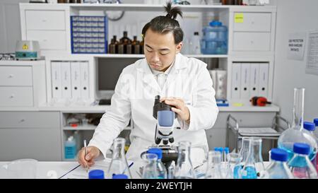 Beau jeune homme scientifique chinois plongeant dans la biologie avec microscope, écrivant des notes d'analyse dans un laboratoire intérieur, entièrement immergé dans la recherche médicale Banque D'Images