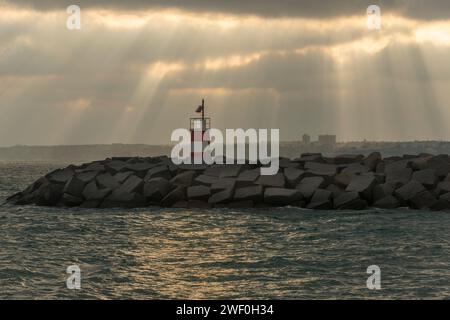 phare rouge et blanc sur des rochers avec ciel orageux et rayons de soleil Banque D'Images