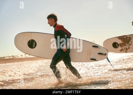 Groupe de surfeurs heureux en combinaison courant dans la mer tenant des planches de surf Banque D'Images