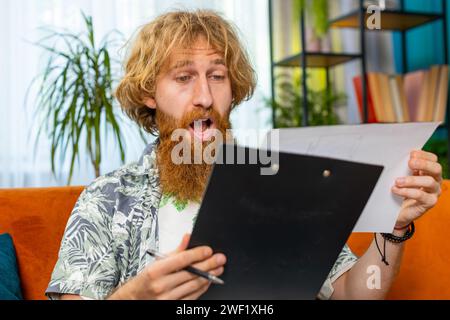 Heureux caucasian Redhead homme freelancer travailler avec des documents de la maison analysant l'information lire des documents d'affaires préparer le rapport financier. Gagnant barbu choqué par la victoire soudaine bonnes nouvelles de succès Banque D'Images