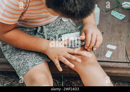 Gros plan d'une blessure à la jambe d'un enfant. L'enfant s'est blessé au genou. Un garçon met un Band-Aid sur une blessure après être tombé sur son genou dans la rue Banque D'Images