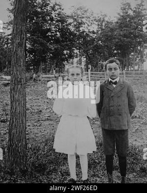 Deux enfants à l'apparence sévère posent debout juste à côté d'un arbre, CA. 1920. Banque D'Images