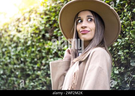 Vue de face d'une jeune femme en veste en cuir noir souriant et tenant son chapeau avec des feuilles vertes en arrière-plan à l'extérieur Banque D'Images