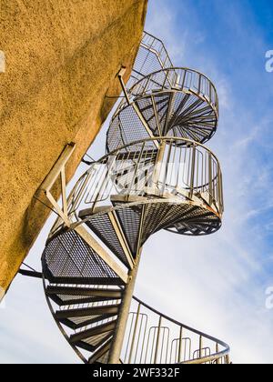 Cette image capture la conception complexe d'un escalier en colimaçon métallique montant le long de l'extérieur d'un bâtiment beige sous un ciel bleu clair. Banque D'Images