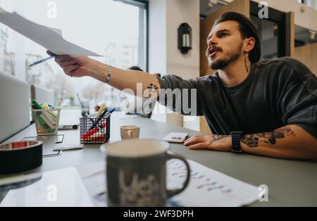 Un jeune professionnel tatoué s’engage activement dans une discussion d’affaires, tenant des documents dans un espace de travail lumineux et contemporain. Banque D'Images