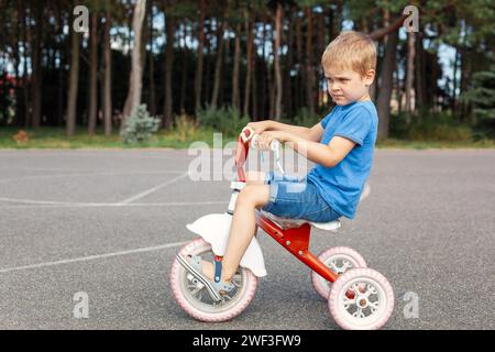 Mignon petit garçon chevauchant un tricycle dans un parc de la ville, photo de profil. Banque D'Images