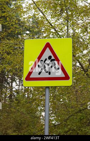 Panneau routier sur poteau, qui est installé près des écoles. Dénotant soyez prudent, les enfants. Carré jaune, triangle rouge, blanc, avec l'image de gens en cours d'exécution. Sécurité, avertissement de trafic, forêt d'automne. Banque D'Images