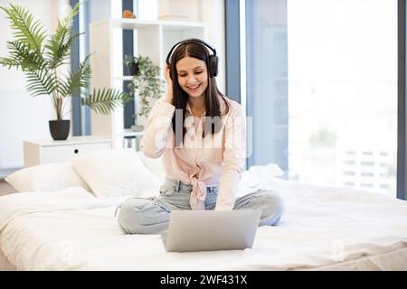 Jeune femme créative travaillant à distance de la maison écoutant de la musique préférée tout en tapant sur un ordinateur portable sans fil moderne. Dame heureuse dans les écouteurs assis sur c Banque D'Images