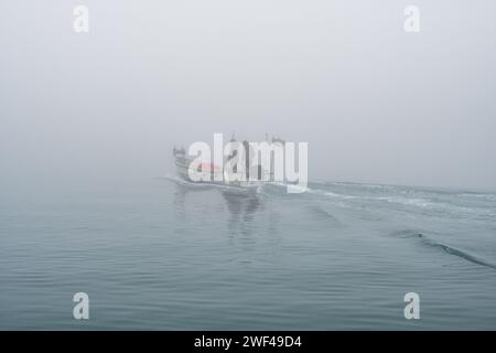 Grado, Italie - 28 janvier 2024 : un petit bateau de pêche avec deux pêcheurs part du port de Mandracchio pour une sortie de pêche par une journée de brouillard épais. Banque D'Images