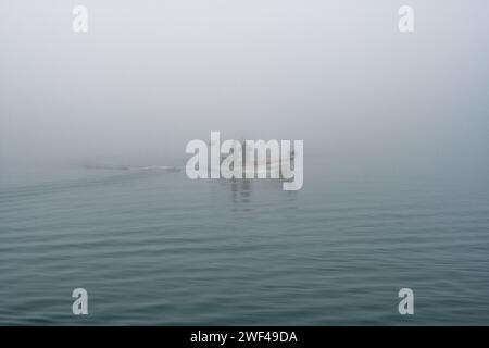 Grado, Italie - 28 janvier 2024 : un petit bateau de pêche avec deux pêcheurs part du port de Mandracchio pour une sortie de pêche par une journée de brouillard épais. Banque D'Images