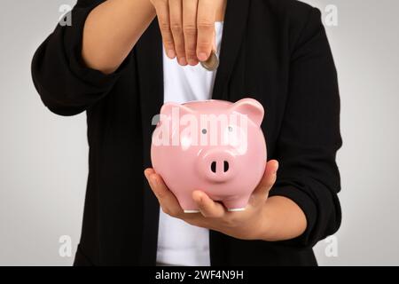 Femme d'affaires habillée intelligemment insérant une pièce de monnaie dans une tirelire rose, illustrant conceptuellement l'épargne Banque D'Images