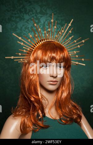 Mannequin de femme aux cheveux rouges en plastique avec de longs cheveux brillants portant une couronne dorée piquante posant sur un fond vert dans un débardeur vert Banque D'Images